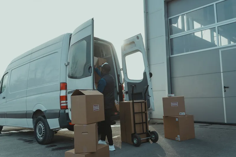 A courier loading taped cardboard parcels into a delivery van outside a warehouse