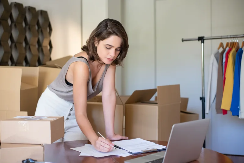 Ecommerce seller writing order details at a desk with a laptop and packed shipping boxes